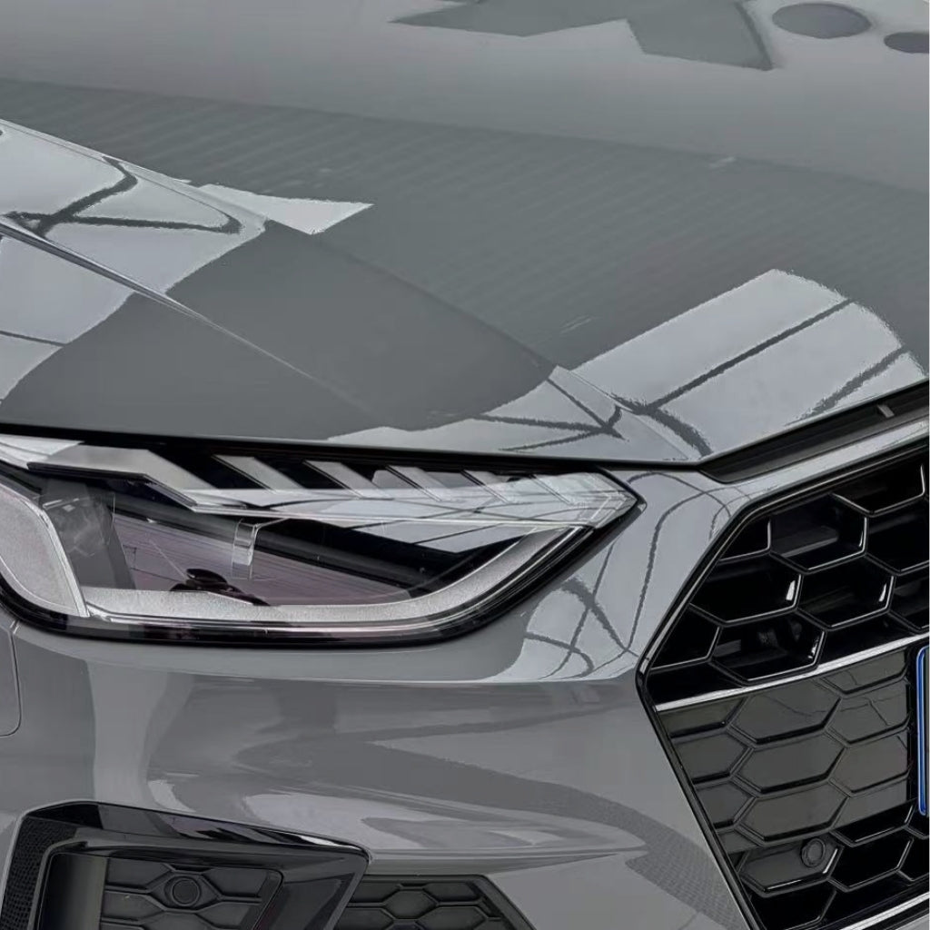 Close-up of the front left headlight and grille of a modern car with Audi Nardo Gray Glossy SelfRepair Nano-Coated PPF, highlighting angular design lines and building reflections on the shiny, protected surface.