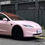 A light pink Tesla Model 3 with black wheels, protected by Baby Pink SelfRepair Nano-Coated PPF for extra scratch protection, is parked near a building with a dark fence and concrete wall.