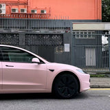 A Baby Pink SelfRepair Nano-Coated PPF car is parked on the street in front of a gray metal fence and an orange building. The photo highlights the car’s front side and wheel.