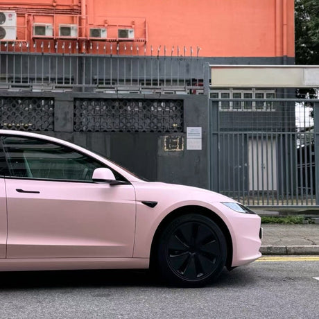 A Baby Pink SelfRepair Nano-Coated PPF car is parked on the street in front of a gray metal fence and an orange building. The photo highlights the car’s front side and wheel.