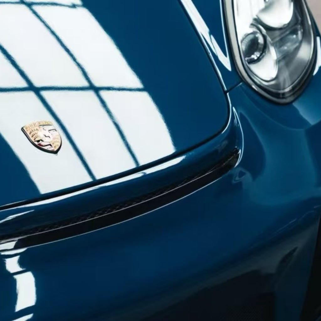 Close-up of a blue Porsche front end with Deep Blue Glossy SelfRepair Nano-Coated PPF protecting the hood and headlight. The Porsche emblem and glossy finish reflect windows, showcasing the quality of this ochranný film na lak.