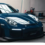 A shiny blue Porsche, shielded by Deep Blue Glossy SelfRepair Nano-Coated PPF, is parked indoors under bright lights with reflections on its hood and windshield. Automotive equipment appears in the background.