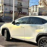 A white SUV with White Diamond Gold Glossy SelfRepair Nano-Coated PPF is parked by apartment buildings on a sunny day, with tree branches and buildings reflected in its tinted windows.