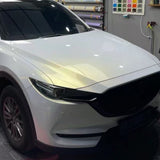 A white SUV is parked indoors by a wall with color samples and rolled-up sheets, featuring a White Diamond Gold Glossy SelfRepair Nano-Coated PPF on the hood and front left side for enhanced protection.