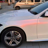 A close-up of a white car's front left side shows its hood protected by White Diamond Pink Glossy SelfRepair Nano-Coated PPF, parked on tiled pavement with sunlight reflecting off the glossy surface; a silver van appears in the background.