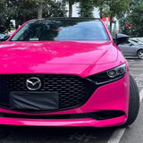 A bright pink Mazda with Hot Crush Glossy SelfRepair Nano-Coated PPF protection is parked in a lot, facing forward. The Mazda logo is visible on the front grille, with trees and other cars in the background.