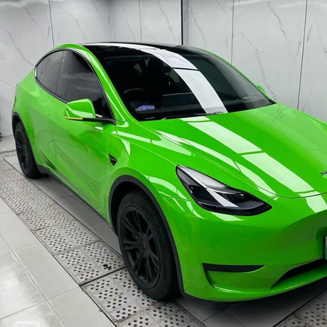 A Tesla Model Y with black rims is parked indoors, featuring Lizard Green Glossy SelfRepair Nano-Coated PPF for body protection, tinted windows, and a black roof, next to a white marble-patterned wall on a tiled floor.