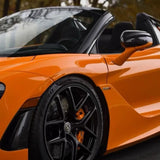 A close-up of a McLaren Orange Glossy sports car with black rims, visible brake calipers, convertible top, and 720S badge. The surface shines with tree and sky reflections, protected by McLaren Orange Glossy SelfRepair Nano-Coated PPF.