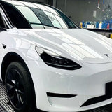 A white Tesla Model Y with black wheels, protected by Metallic White Glossy SelfRepair Nano-Coated PPF, is parked indoors. Window reflections shine on its hood, and shelves of bottles are visible in the background.