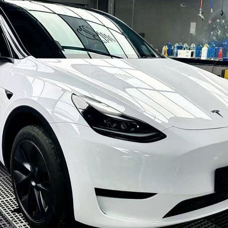 A white Tesla Model Y with black wheels, protected by Metallic White Glossy SelfRepair Nano-Coated PPF, is parked indoors. Window reflections shine on its hood, and shelves of bottles are visible in the background.