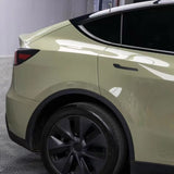 The image shows the rear side of a Tesla Model Y with Military Green Glossy SelfRepair Nano-Coated PPF, highlighting the back door, rear wheel, and tail light with black trim and tinted windows, parked indoors on a patterned floor.