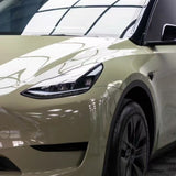 A military green Tesla Model Y seen from the front left, with its headlight, front fender, and part of the hood protected by Military Green Glossy SelfRepair Nano-Coated PPF, reflecting a grid-patterned ceiling above.
