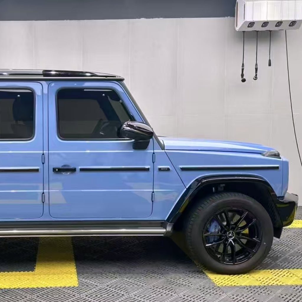 A blue Mercedes-Benz G-Class SUV, protected by Porcelain Blue Glossy SelfRepair Nano-Coated PPF for dokonalá ochrana laku a samoregeneračná technológia, is parked indoors on a yellow-striped floor with a gray wall and AC unit in the background. Only the left side and front wheel are visible.