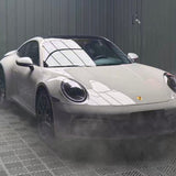 A white Porsche sports car is parked indoors on a patterned floor, mist swirling by. Its glossy reflection shines—protected by Porsche Gray Glossy SelfRepair Nano-Coated PPF for top-tier paint protection and self-healing performance.