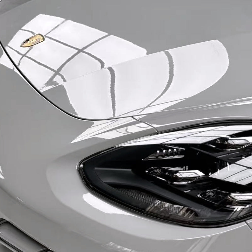 Close-up of the front left headlight and hood of a white Porsche with Porsche Gray Glossy SelfRepair Nano-Coated PPF, featuring a shiny surface and window grid reflections for superior paint protection.