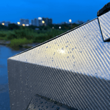 Close-up of Liquid Metallic Aluminium Carbon Fiber Self-Healing Nano-Coated PPF with water droplets at dusk. Blurred city lights and a blue sky in the background highlight its sleek, metallic carbon fiber finish.