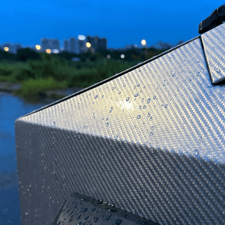 Close-up of Liquid Metallic Aluminium Carbon Fiber Self-Healing Nano-Coated PPF with water droplets at dusk. Blurred city lights and a blue sky in the background highlight its sleek, metallic carbon fiber finish.