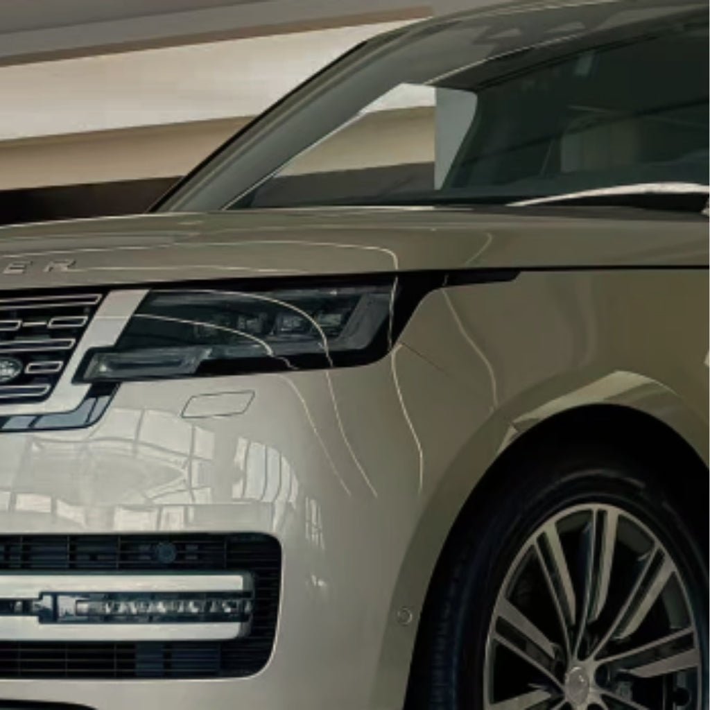 Close-up of the front left side of a beige luxury SUV with headlight, grille, and large alloy wheel, protected by Batumi Gold Liquid Metallic Glossy SelfRepair Nano-Coated PPF. The vehicle is parked indoors under bright lighting.