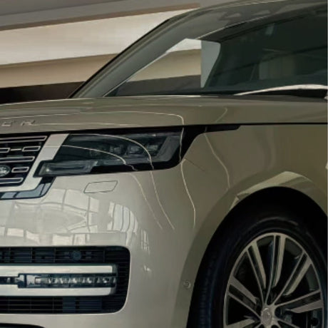 Close-up of the front left side of a beige luxury SUV with headlight, grille, and large alloy wheel, protected by Batumi Gold Liquid Metallic Glossy SelfRepair Nano-Coated PPF. The vehicle is parked indoors under bright lighting.