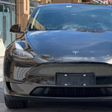 A close-up front view of a black Tesla with Diamond Black Gold Glossy SelfRepair Nano-Coated Nexthor™ PPF, highlighting its grille, headlights, Tesla logo, and building reflections on the hood and windshield.