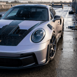 A high-performance sports car with a matte pink and black, carbon-inspired finish features Forged Carbon Graphite Self-Healing Nano-Coated PPF, aerodynamic vents, racing wheels, and a large rear wing on a wet racetrack.