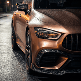 Close-up of the front left side of a bronze sports car with raindrops, protected by Forged Carbon Graphite Self-Healing Nano-Coated PPF, parked on a wet street at night under dim streetlights.