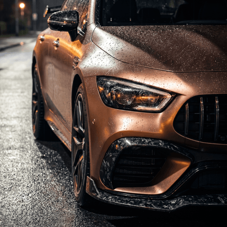 Close-up of the front left side of a bronze sports car with raindrops, protected by Forged Carbon Graphite Self-Healing Nano-Coated PPF, parked on a wet street at night under dim streetlights.