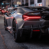 A sports car protected by Forged Carbon Graphite Self-Healing Nano-Coated PPF, featuring a matte gray carbon finish and glowing red taillights, is parked on a city street at night amid buildings and traffic lights.