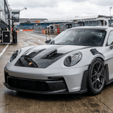 A silver sports car with black accents, protected by Forged Carbon Graphite Self-Healing Nano-Coated PPF, is parked on a wet race track near pit garages. Its low, aggressive stance and large wheels stand out against the cloudy racing circuit backdrop.