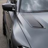 Close-up of the front left side of a sleek car featuring Matte Carbon Fiber SelfRepair Nano-Coated PPF, displaying the hood, side mirror, and carbon fiber vent detail with cloudy sky reflections on its matte surface.
