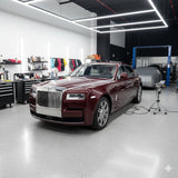 A shiny maroon Rolls-Royce sedan, shielded by Diamond Black Rose Glossy SelfRepair Nano-Coated PPF, is parked in a modern garage with bright lights, tools, and detailing supplies on the walls. Another car sits covered in the background.