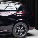 A close-up side view of a Nexthor dark maroon BMW SUV with Magic Black to Red Charm Glossy SelfRepair Nano-Coated PPF, focusing on the rear door, window, and wheel. Reflections gleam on the surface, set against black curtains and a white floor.