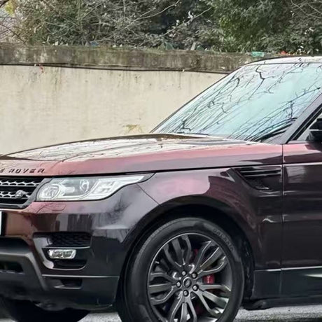 A Range Rover SUV with Magic Black to Red Charm Glossy SelfRepair Nano-Coated PPF is parked near a concrete wall and trees, showing its front and side, distinctive grille, and alloy wheels.