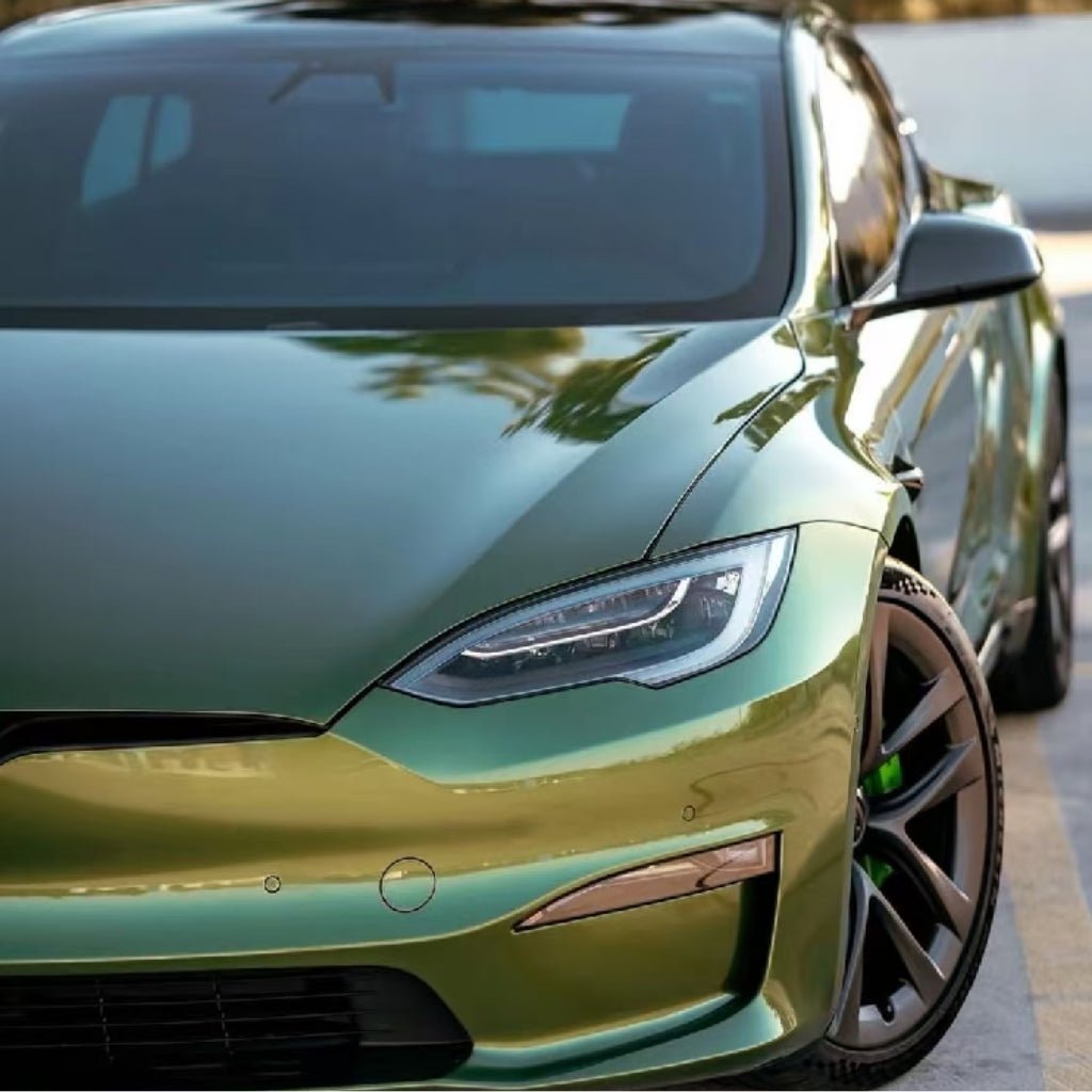 Close-up of a green metallic Tesla Model S outdoors, featuring sleek front design and sharp headlights, protected by Blue Gold Green Chameleon SelfRepair Nano-Coated PPF that keeps the paint flawless and reflective with trees mirrored on the hood.