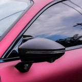 Close-up of a black side mirror on a matte pink car wrapped with Diamond Brilliant Magenta Glossy SelfRepair Nano-Coated PPF, featuring tinted windows and a camera under the mirror, reflecting a blue sky with clouds.