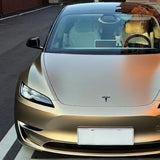 A gold Tesla with Champagne Gold Liquid Matte Metallic SelfRepair Nano-Coated PPF is parked on the street. Plush toys and a small figure decorate its dashboard, visible through the windshield, and the license plate is blank.