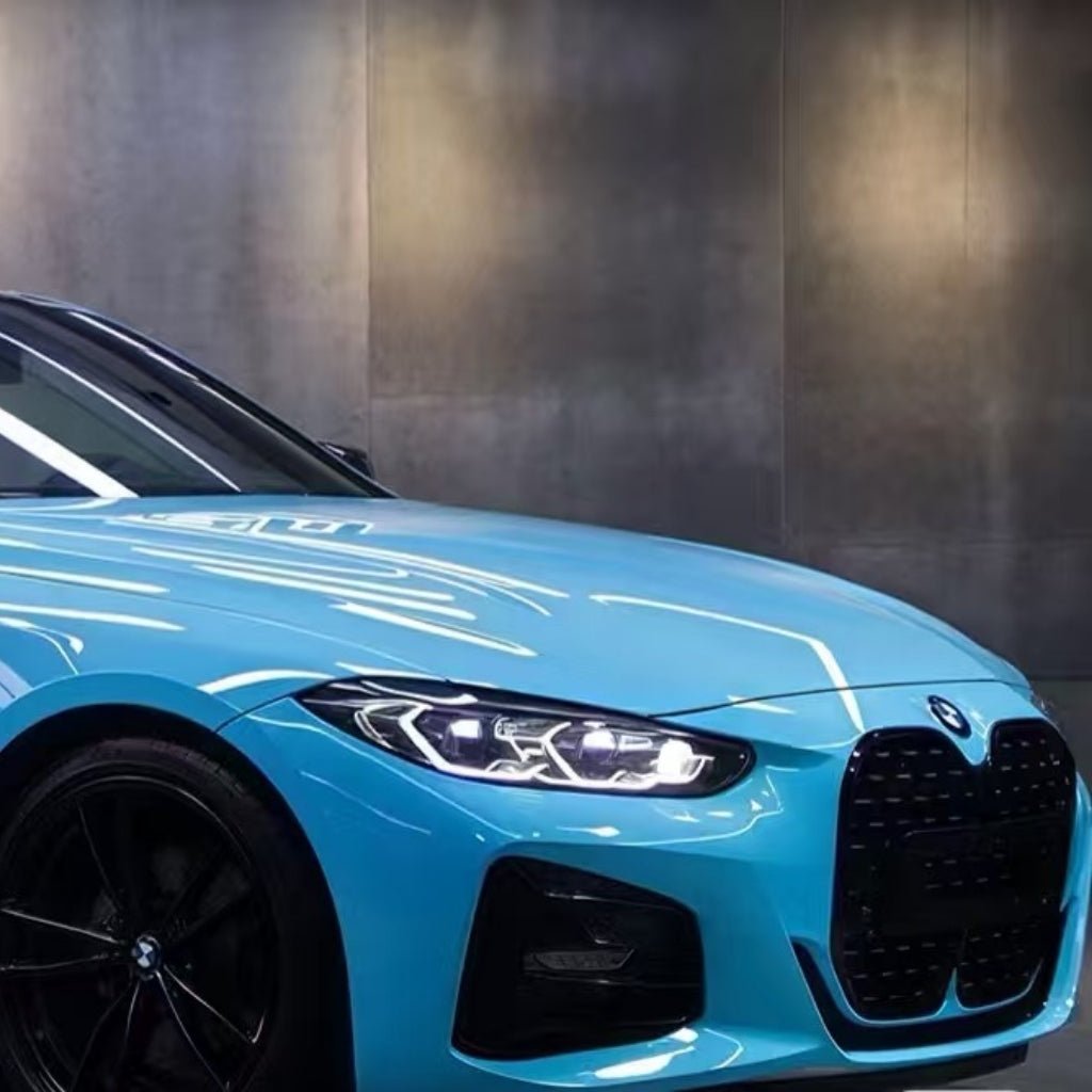 A close-up of the front left side of a blue BMW, featuring its illuminated headlight, black grille, and sleek design protected by Sky Blue Charm SelfRepair Nano-Coated PPF against a modern gray concrete wall.
