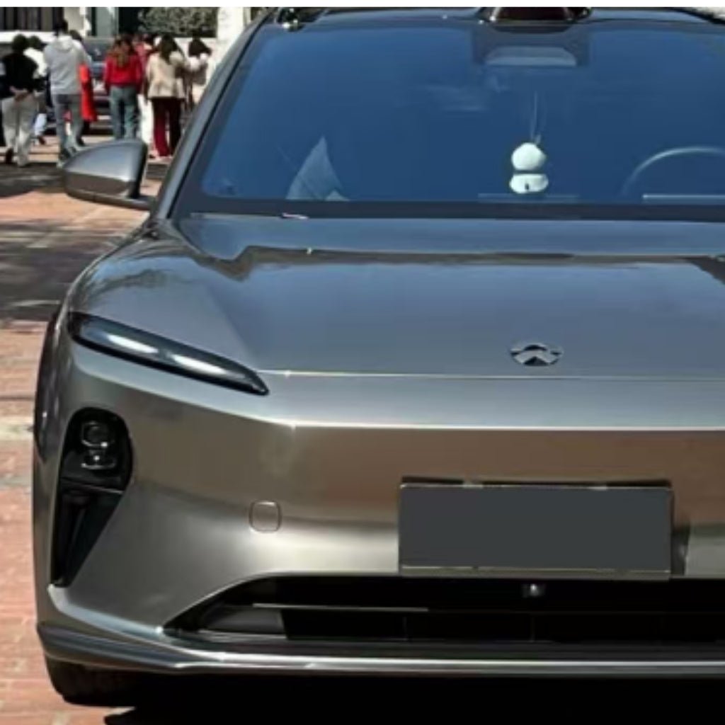 A sleek metallic gray electric car is parked outdoors, protected by Chrome Black Liquid Metallic Glossy SelfRepair Nano-Coated PPF. The NIO logo is on the front grille, and a small mirror decoration hangs inside as people walk in the blurred background.