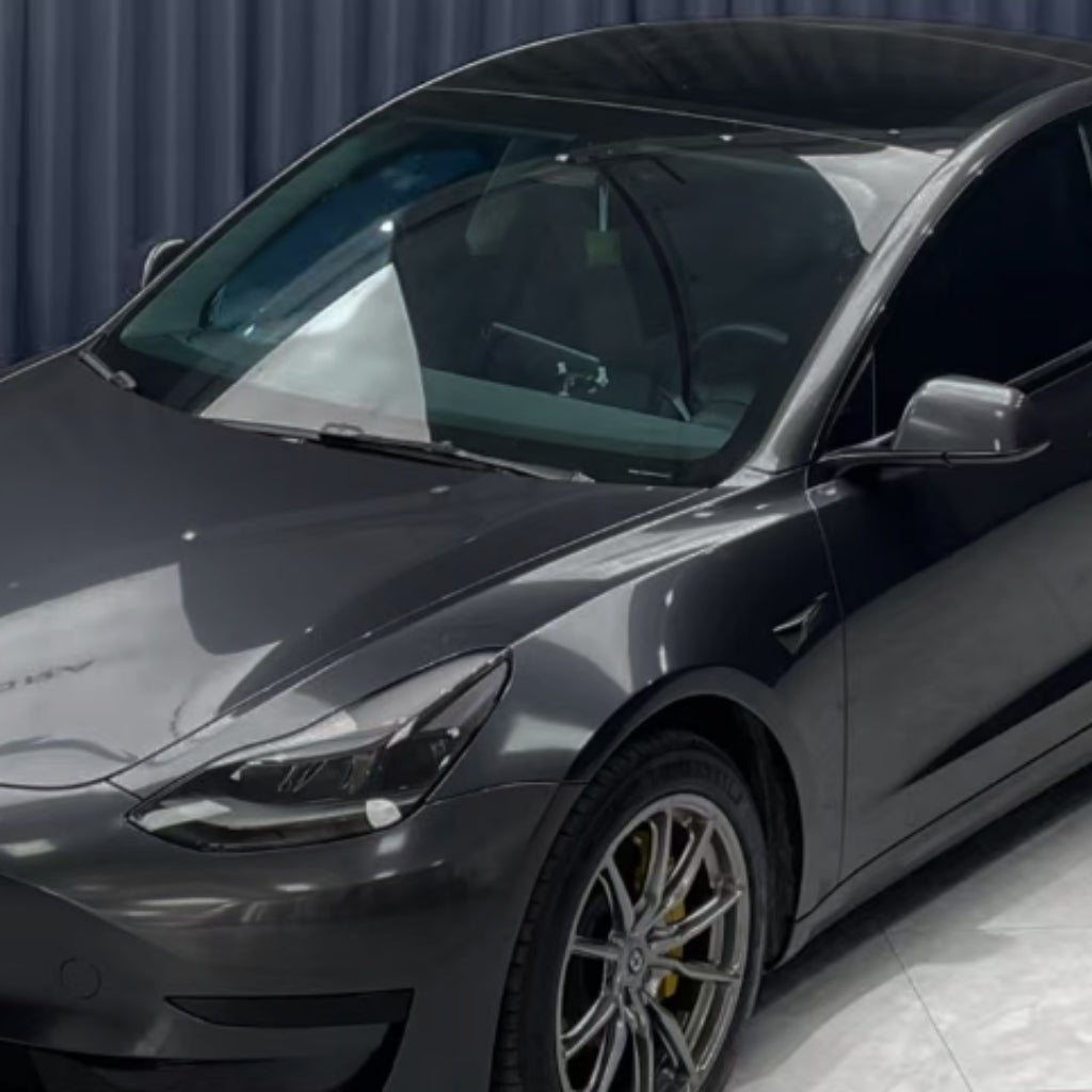 A sleek, dark grey Tesla Model 3 in Diamond Coal Gray Glossy SelfRepair Nano-Coated PPF is parked indoors on a shiny floor with a dark curtain backdrop. The futuristic design, tinted windows, and advanced protective PPF film stand out.