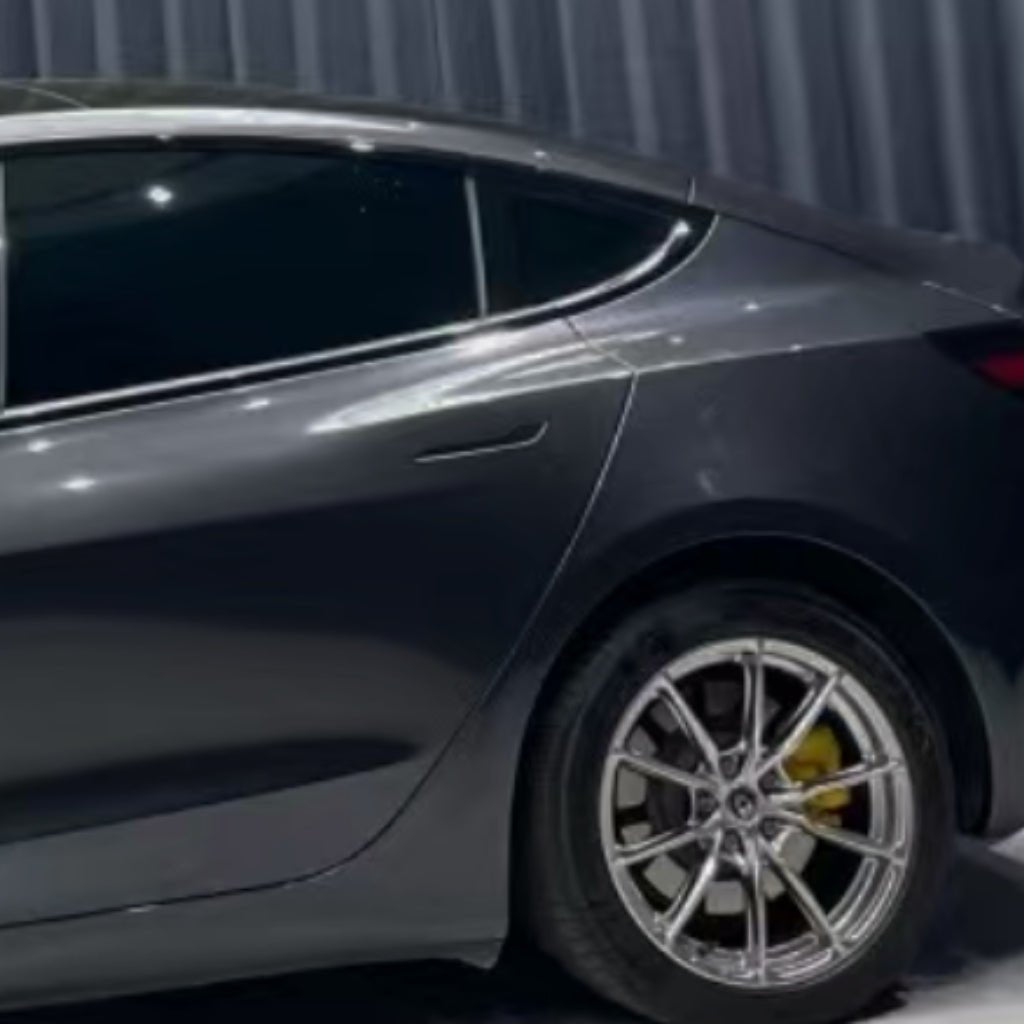 A close-up side view of a car wrapped in Diamond Coal Gray Glossy SelfRepair Nano-Coated PPF, highlighting the rear door, tinted window, silver alloy wheel with yellow brake caliper, and a corrugated metal wall in the background.
