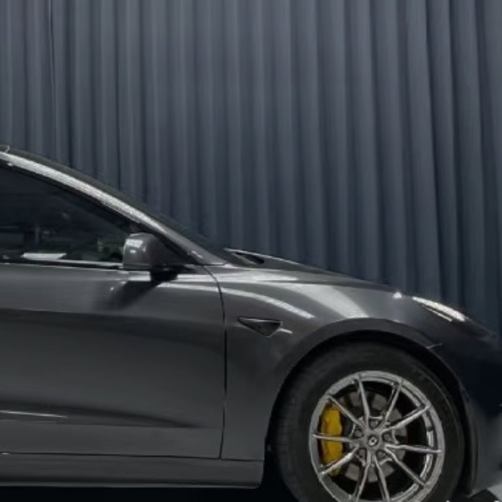 A dark gray car with yellow brake calipers, protected by Diamond Coal Gray Glossy SelfRepair Nano-Coated PPF, is parked in front of vertical black curtains, showing its front driver’s side and part of the windshield.