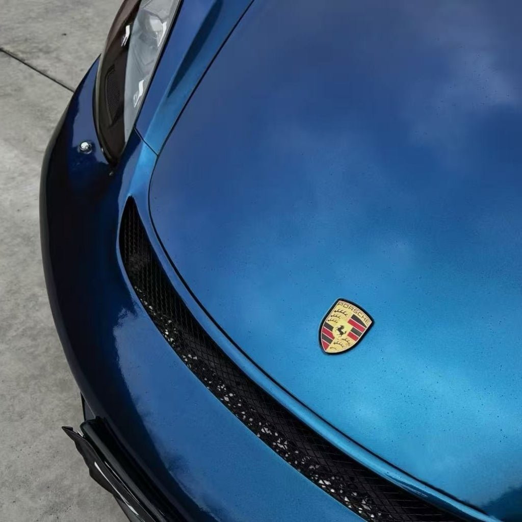 Close-up of a blue Porsche hood protected by Diamond Dark Shadow Blue Glossy SelfRepair Nano-Coated PPF, featuring the Porsche logo and cloud reflections. The angled view highlights the glossy finish and advanced self-repair nanotechnology film.