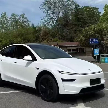 A white Tesla featuring tinted windows, black wheels, and White Diamond Glossy SelfRepair Nano-Coated PPF is parked in a lot by trees and a small building, with a blue exit sign visible in the background.