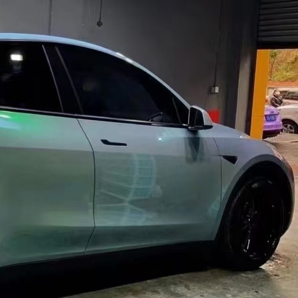 A shiny, light-colored car with tinted windows and black wheels features Gray to Green Charm Glossy SelfRepair Nano-Coated PPF, parked indoors near a partially open garage door with another car and scooter visible outside.