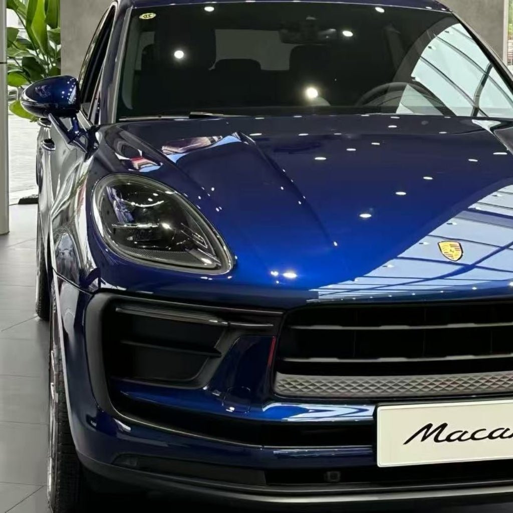 A close-up front view of a Porsche Macan wrapped in Diamond Gentian Blue Glossy SelfRepair Nano-Coated PPF, parked indoors and highlighting its sleek design, headlights, partial grille, and the Porsche emblem on the hood.