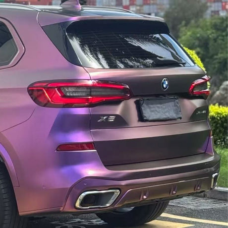A BMW X5 with a shiny iridescent purple wrap, featuring Gold White Red Chameleon SelfRepair Nano-Coated PPF, is parked by the curb. Sunlight reflects off its surface. The license plate is blurred.