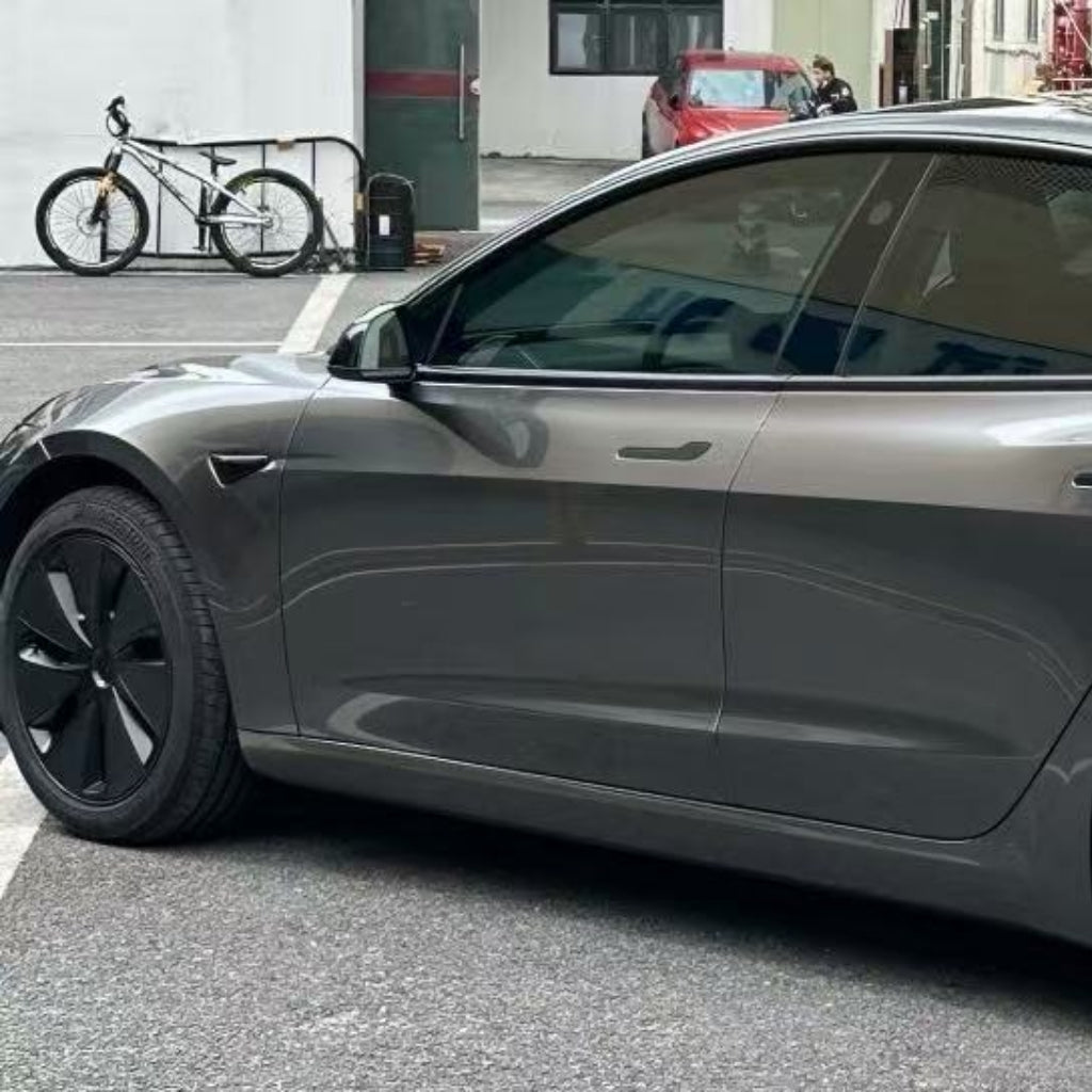 A gray Tesla Model 3 featuring Gray Liquid Metallic Glossy SelfRepair Nano-Coated PPF is parked on a city street beside a locked bicycle. Nearby buildings and a red car accentuate its sleek appearance and advanced nano-coated protection film.