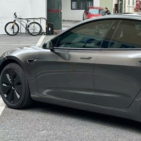 A gray Tesla Model 3 featuring Gray Liquid Metallic Glossy SelfRepair Nano-Coated PPF is parked on a city street beside a locked bicycle. Nearby buildings and a red car accentuate its sleek appearance and advanced nano-coated protection film.