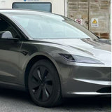 A sleek, silver electric car with black wheels and minimalist headlights is parked on the street, protected by Gray Liquid Metallic Glossy SelfRepair Nano-Coated PPF. In the background, a building displays warning signs and closed shutters.