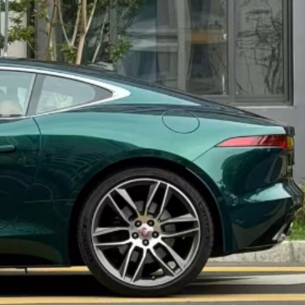 A close-up side view of a green sports car's rear with sleek lines and red taillights, featuring Diamond British Green Glossy SelfRepair Nano-Coated PPF, parked by a sidewalk with greenery and building reflections.